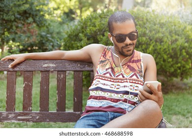 African guy in earphones sitting on bench in city park listening to music on his smartphone, checking e-mail using Internet-enabled mobile phone, liking posts and leaving comments on social networks