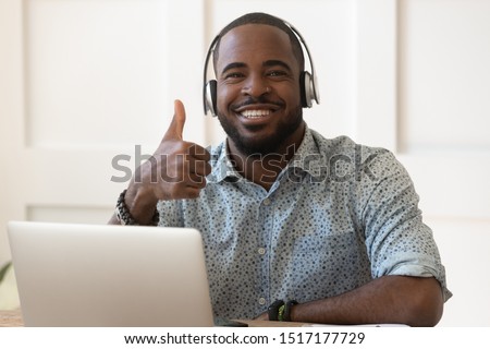 African guy in headphones sitting near computer smiling looking at camera showing thumbs up, online audio educational course advertisement, teacher and student successful distant communication concept