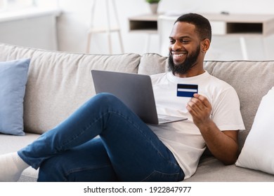 African Guy Holding Credit Card Shopping Online Via Laptop Computer Sitting On Couch At Home. Happy Customer Buying Clothes Concept. Shopaholism And E-Commerce. Selective Focus