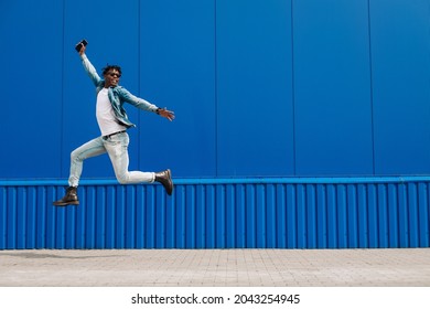 African Guy Jumping With A Phone In His Hands, In The City Against The Background Of A Blue Building, Concept Of Success