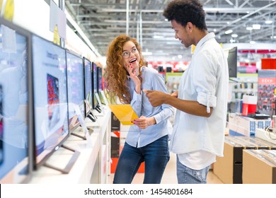 African Guy With Red-haired Girl Standing In Front Of The Stand In The Electronics Store Choose A Plasma TV Looking At The Price Tags