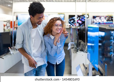 African Guy With Red-haired Girl Standing In Front Of The Stand In The Electronics Store Choose A Large Plasma TV Before Buying