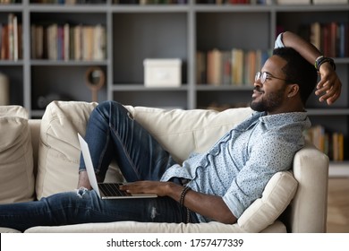 African Guy Relieving Fatigue Resting On Comfy Couch In Modern Living Room Put On Lap Notebook Using Media Services App Distracted From Gadget Looks At Window Daydreaming Feels Good No Stress Concept