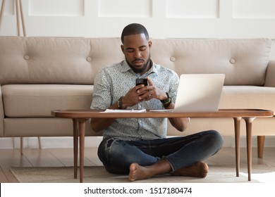 African guy sits leaned on couch cross-legged on carpet near coffee table hold cellphone distracted from work chat online, self-employed man solve business issues contact with client distantly concept
