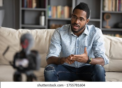 African guy sitting on couch filming online training webinar using camcorder dslr modern professional device camera. Influencer recording advertisement feedback for client, makes job interview concept