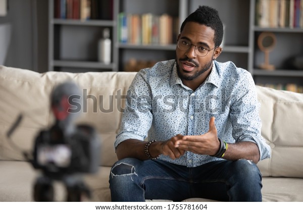 African guy sitting on couch filming online\
training webinar using camcorder dslr modern professional device\
camera. Influencer recording advertisement feedback for client,\
makes job interview\
concept