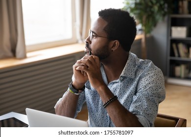 African guy sitting at table near laptop pondering over problem search answer looking at distance out the window. Student or office worker having doubts, feel uncertain thinking about solution concept