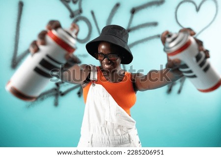 African Hipster girl painting graffiti on teal wall holding spray bottles wearing orange sunglasses 