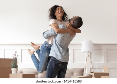 African Husband Lifting Happy Wife Laughing Celebrating Moving Day With Boxes, Excited First Time Home Buyers Renters Owners Having Fun Enjoy Relocation Into Own New Flat House, Mortgage Investment