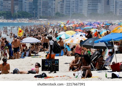 African Immigrant Vendor Walking At Copacabana Beach. Black Man Selling Goods To Beachgoers Near Shore Among People Sunbathing On Colorful Umbrellas - Rio De Janeiro, Brazil 12.31.2020