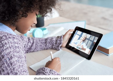 African Kid Child Girl Holding Digital Tablet Talking With Remote Teacher Tutor On Zoom Distance Video Conference Call Elearning Online Virtual Class. Children Learning At Home, Over Shoulder View.