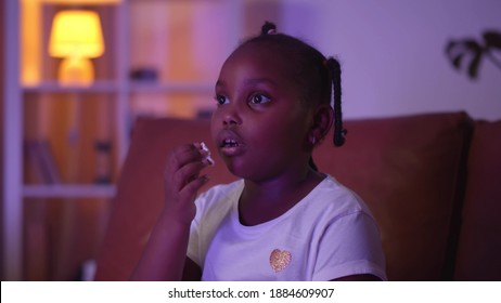 African Little Girl Watching Tv And Eating Popcorn In Evening At Home. Portrait Of Cute Preteen Afro-american Child Enjoying Cartoon And Eating Popcorn Relaxing On Sofa In Dark Living Room