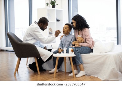 African male doctor examines young African boy's throat with wooden spatula. Boy sits on bed in hospital room with concerned mother holding teddy bear. - Powered by Shutterstock