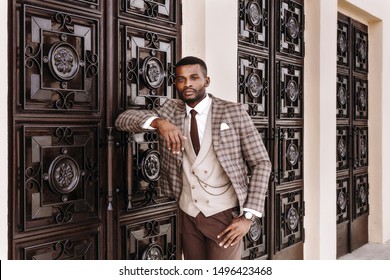 african male model in a brown suit posing outside, leaning on the decorated wall, entrance door, stylish luxury expensive business look, well dressed gentleman, urban men, city life, high fashion look