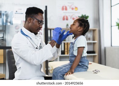 African male physician in uniform and gloves checking little girl's throat at modern clinic. Pretty female patient say aaah during check up at hospital. - Powered by Shutterstock