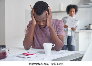 African Male Sitting At Kitchen Table, Feeling Sad And Depressed About Debts, Can't Pay Off His Utility Bills. Wife Standing In Background With Papers From Bank. People And Finances Concept