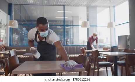 African male worker cleaning table with disinfectant in restaurant during coronavirus outbreak. Waiter in protective mask and gloves disinfecting table with spray and cloth