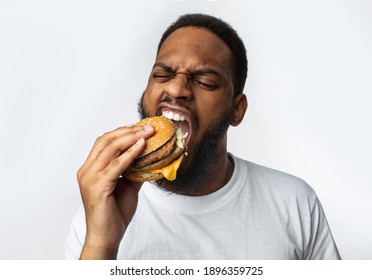 African Man Biting Burger Enjoying Unhealthy Tasty Junk Food Standing In Studio Over White Background. Overeating, Fast Food And Male Nutrition Concept. Funny Hungry Black Guy Eats Cheeseburger