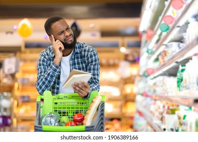 African Man Buying Food With Groceries Checklist Standing With Cart Near Shelf In Supermarket. Grocery Shop Customer Calculating Increased Prices. Consumerism And Crisis. Blank Space For Text