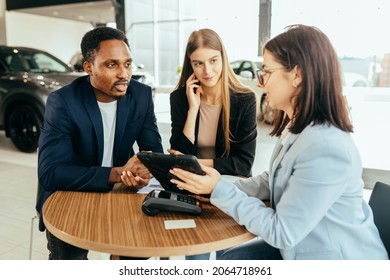 African Man And Caucasian Woman Talking With Female Car Dealer At Auto Salon While Making Expensive Purchase. Couple Choosing New Vehicle Together.