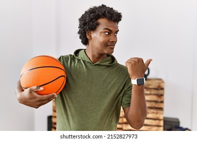 African man with curly hair holding basketball ball at the gym pointing thumb up to the side smiling happy with open mouth 