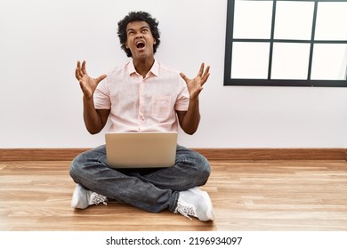 African Man With Curly Hair Using Laptop Sitting On The Floor Crazy And Mad Shouting And Yelling With Aggressive Expression And Arms Raised. Frustration Concept. 