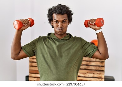 African man with curly hair wearing sportswear using dumbbells puffing cheeks with funny face. mouth inflated with air, catching air. 