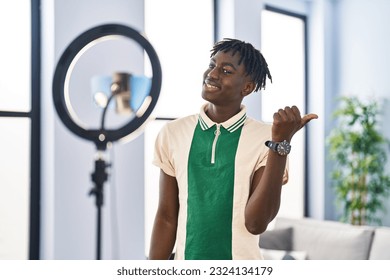 African man with dreadlocks recording vlog tutorial with smartphone at home pointing thumb up to the side smiling happy with open mouth 