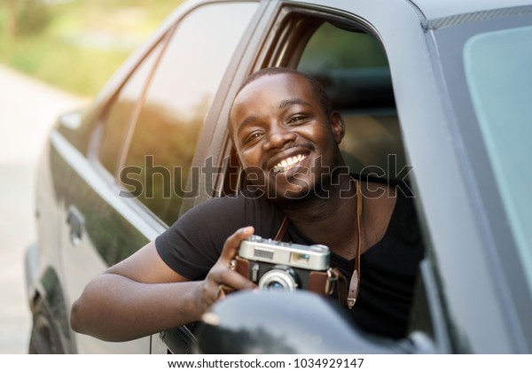 African man driver holding a film\
camera and smiling while sitting in a car with open front\
window.