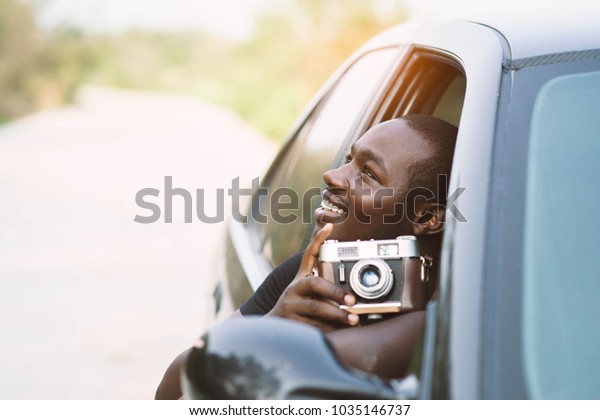African man driver holding a film
camera and smiling while sitting in a car with open front
window.