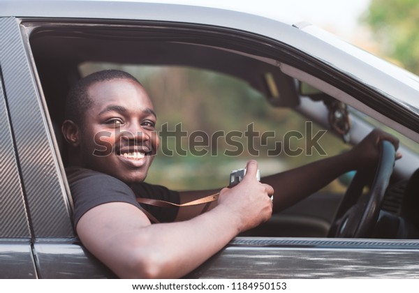 African man driver holding a film\
camera and smiling while sitting in a car with open front\
window.