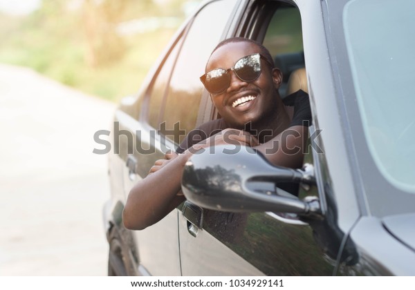 African man driver Wearing\
sunglasses and smiling while sitting in a car with open front\
window.