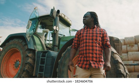 African Man, Farmer Standing In Front Of The Green Tractor . Hay Rools Stack In The Background. High Quality Photo