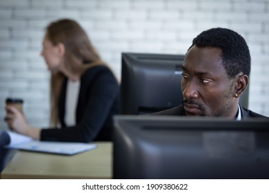 An African Man Looked Around The Office With A Serious Expression. A Male Office Worker Paranoid His Coworker As He Is Secretly Doing Something On The Desktop. 