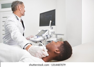 African Man Lying In Medical Cabinet. Sonography, Ultrasound Diagnostics Of Lymph Nodes On Neck. Doctor Wearing In White, Medical Gown And Gloves Doing Examination With Ultrasound Probe.