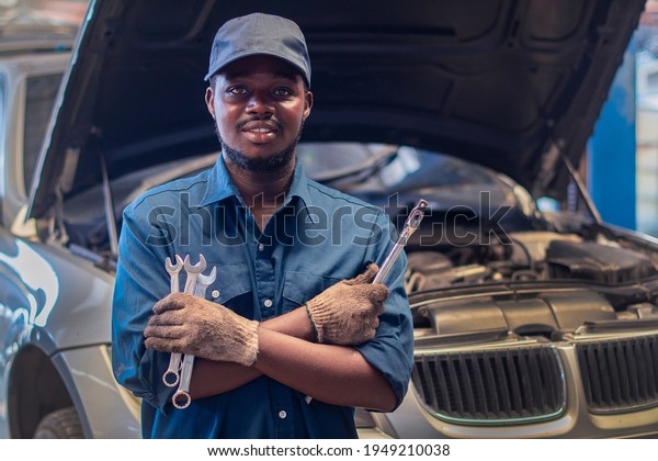 African man mechanic in uniform\
with crossed arms and wrenches standing at the car repair\
station