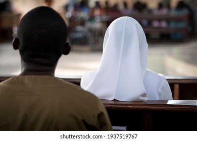 An African Man And A Nun Dressed In White Sit On Wooden Church Pews For A Catholic Mass In Ghana, West Africa.