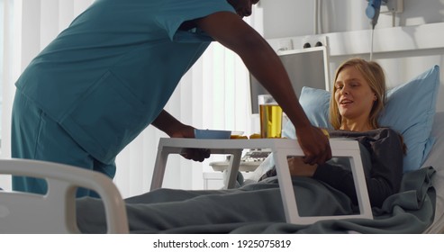 African Man Nurse Serving Food In Hospital To Patient In Bed. Male Medical Worker Serving Young Female Patient Meal In Hospital Bed. Healthcare And Medicine Concept