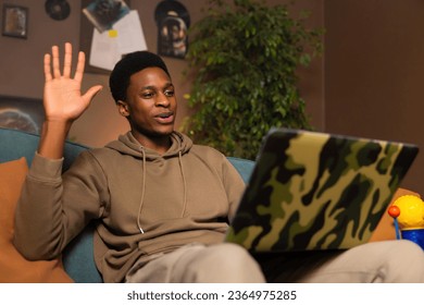African man on cozy couch in a well-lit living room, waving at the camera on his laptop engaging with his device communicating with friends or family members through video chat or social media. - Shutterstock ID 2364975285