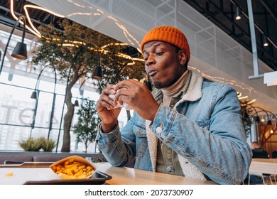 African Man In An Orange Hat And Denim Jacket Eats A Delicious Burger And French Fries In A Shopping Mall On A Food Court, Greasy And Unhealthy Fast Food