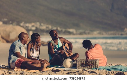 African Man Playing Guitar For Friends On The Beach. Group Of Friends Having Picnic At The Seashore.