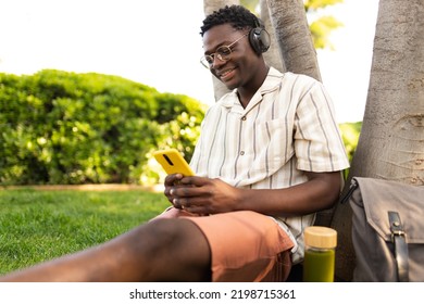 African Man Relaxing Outdoors Listens To Music Using Phone And Headphones. Black College Student On Campus. Copy Space