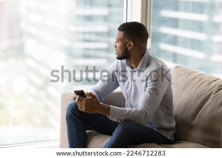 African man resting on couch holds smart phone staring out window at big city view, enjoy cityscape using modern technology. Lifestyle, wireless tech usage, user of e-services for comfort urban life