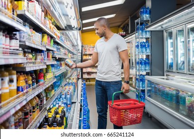 African Man Shopping In Beverage Section At Supermarket. Black Man Doing Shopping At Market While Buying Cold Drink. Handsome Guy Holding Shopping Basket Reading Nutritional Values Of Product.