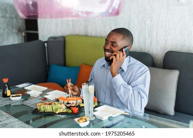 African Man Sits In A Restaurant, Talking On The Phone And Eating Japanese Sushi. Smiling Happy Business Man Eats Sushi With Chopsticks And Talks With Friend During Lunch Time