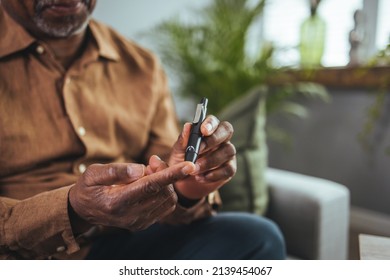 African Man Sits At A Table In The Living Room And Takes Blood From His Finger Due To Diabetes. Everyday Life Of A Man Of African American Nationality With A Chronic Disease Who Uses A Glucose Tester.