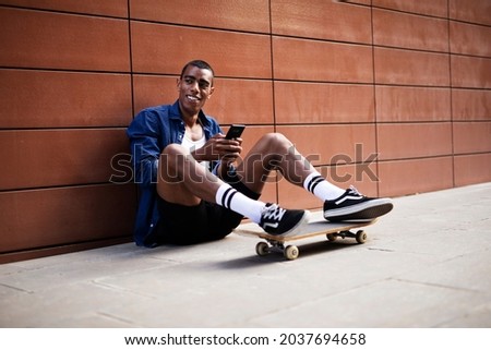 African man with skateboard. Young handsome man sitting on the floor uisng the phone