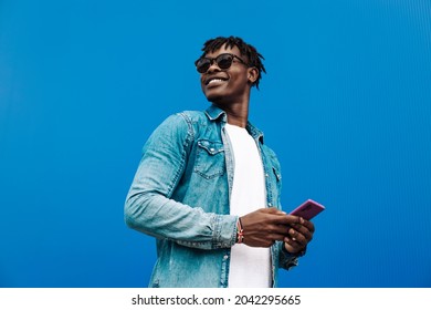 African Man Typing Sms On Blue Background, 5g Internet Concept, High Speed Internet On Phone, Black Man In Blue Sacker, And Jeans