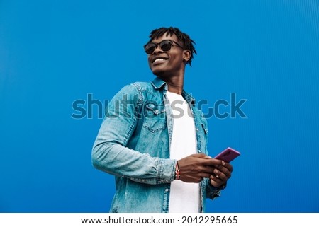 african man typing sms on blue background, 5g internet concept, high speed internet on phone, black man in blue sacker, and jeans