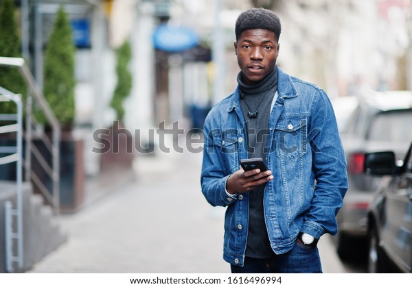 African man wear on jeans\
jacket posed outdoor, standing against black car with mobile phone\
at hand.